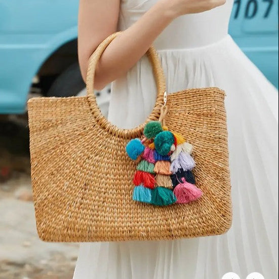 Woven handbag with colorful pom-poms held by a person in a white dress, with a blurred background.